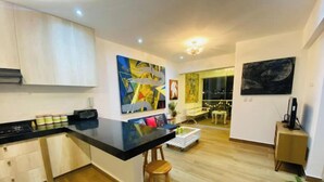 A modern kitchen with a black countertop, wooden cabinets, and a stool. There are two paintings on the wall and a potted plant on the floor. - Beach Apartment /Gallery in Punta Hermosa (Lima)