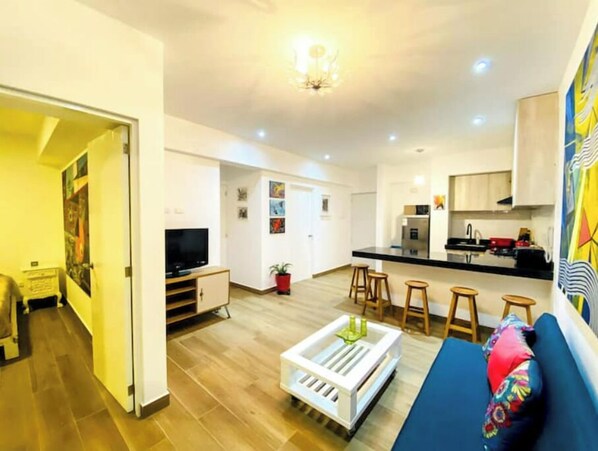 Ein modernes Wohnzimmer mit Sofa, Couchtisch und Fernseher. Es gibt eine Küchenzeile mit Kühlschrank, Herd und Barhocker. Der Raum hat Holzböden und weiße Wände.