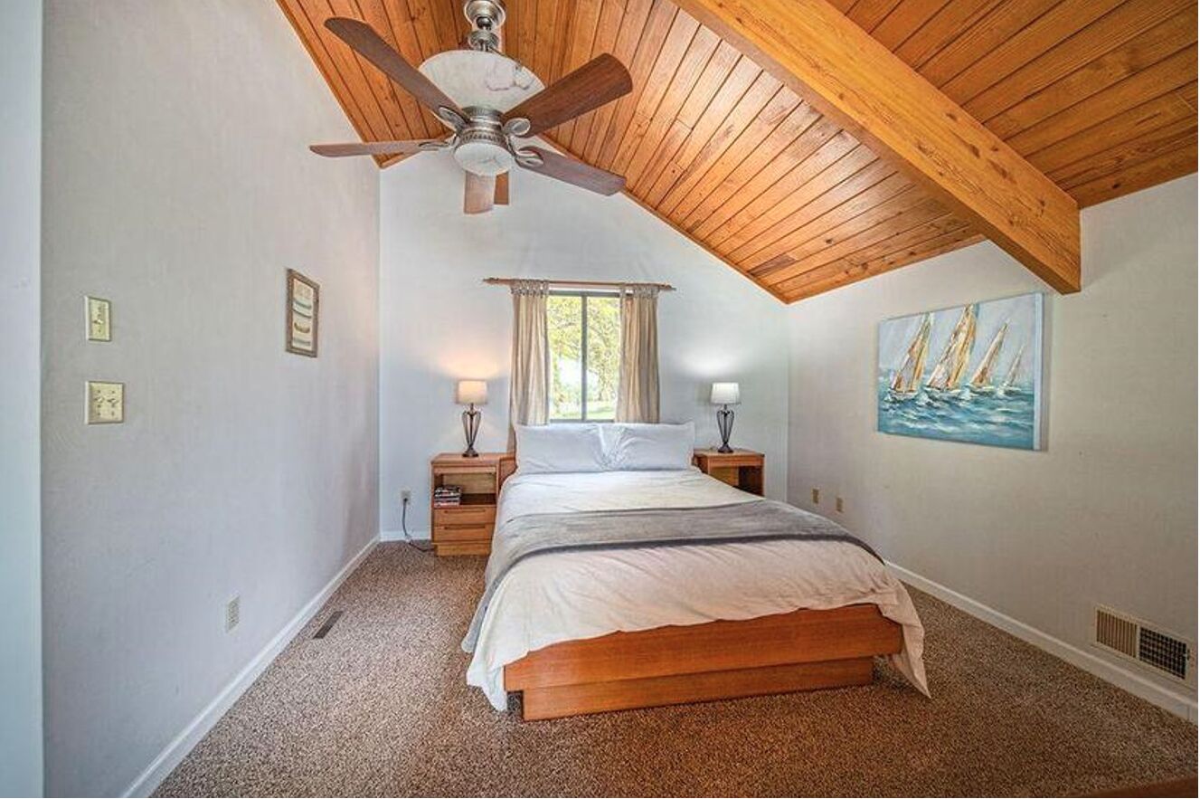 Quarto com teto de madeira, uma cama, duas mesas de cabeceira, uma janela com cortinas e um quadro na parede.