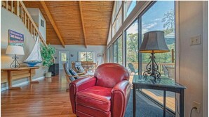 Una habitación amplia con techo de madera, un sillón de cuero rojo, una mesa con lámpara y vistas al exterior a través de grandes ventanales.