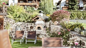 A stone-walled outdoor seating area with wooden chairs and a table, surrounded by lush greenery and flowering plants. - Centrally located, family-friendly apartment in the middle of the Dolomites (Sankt Ulrich)