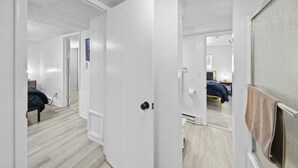 Un pasillo con una cama, una puerta y un espejo que refleja una habitación con cama y ventana.