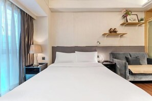 A hotel room with a large bed, a grey sofa, a nightstand with a lamp, and a wall-mounted shelf with decorative items.