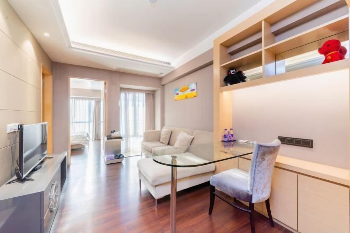 A modern living room with a flat-screen TV, a glass coffee table, a sofa, and a dining area with a glass table and chairs.