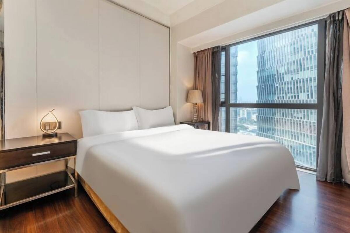 Une chambre d’hôtel moderne avec un grand lit, une table de chevet, une lampe et une vue sur le paysage urbain, visible à travers une grande fenêtre.