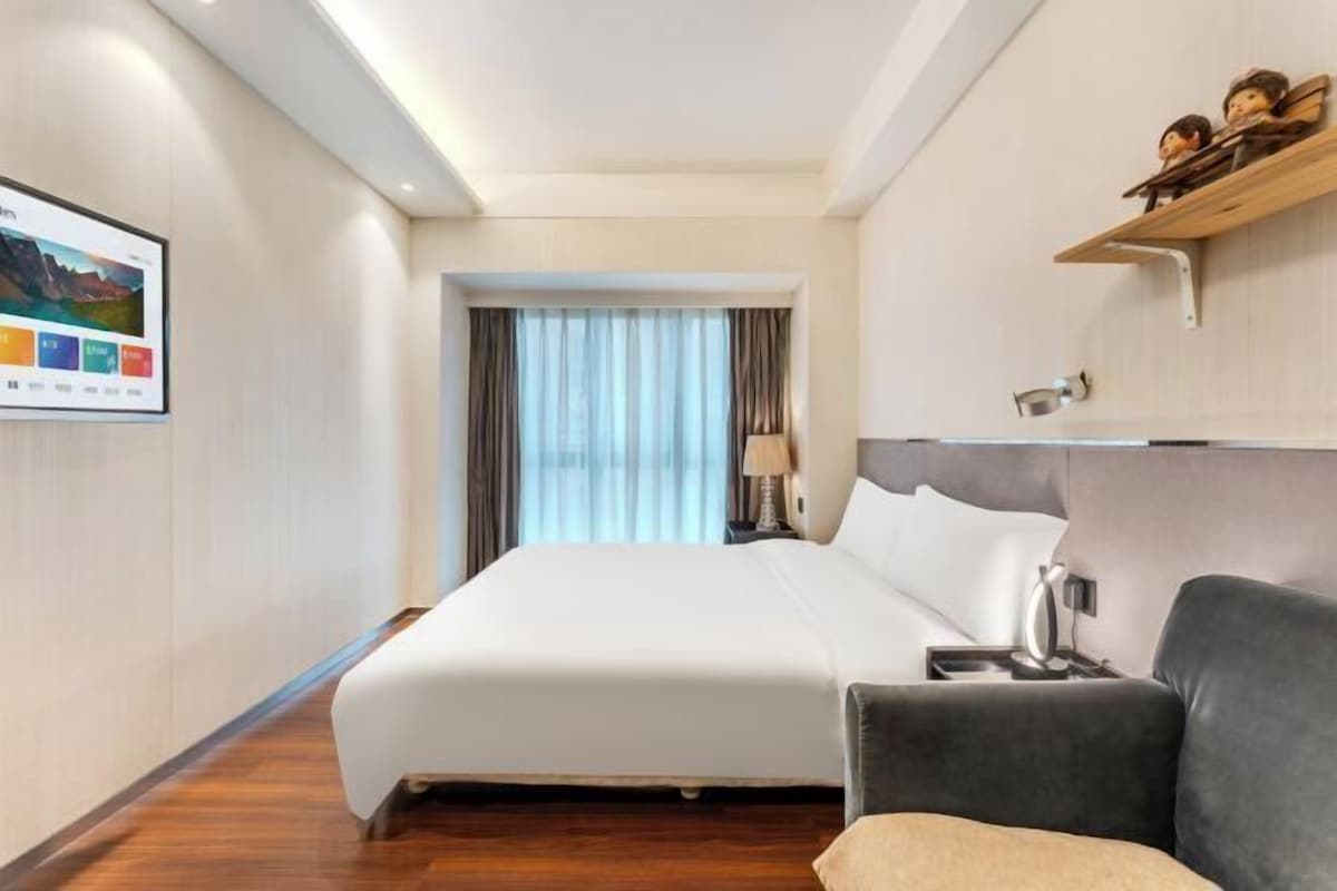 A hotel room with a large bed, a sofa, a desk with a lamp, and a wall-mounted shelf with decorative items.