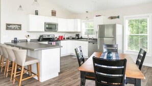 A modern kitchen with white cabinets, a central island with bar stools, a dining table with chairs, and a refrigerator.