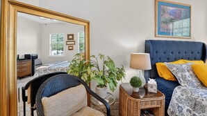 Une chambre avec un grand miroir, un lit avec une literie bleue et jaune, une chaise en osier, une table de chevet avec une lampe et une plante en pot.