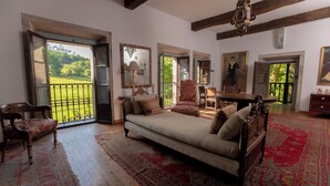 A spacious room with a large bed, a wooden chair, and a dining table. There are multiple windows providing views of a garden.