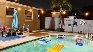 A swimming pool with inflatable toys, a covered patio with chairs, and string lights. - Fun Kids Family Stay - FLL Cruise Guests & Airport! AirHockey-Arcade-Pool-Toys (Hollywood)