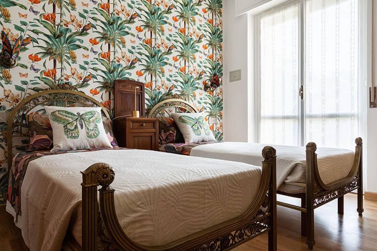 Une chambre à coucher avec deux lits en bois, un papier peint à motifs floraux et une fenêtre ornée de rideaux légers.