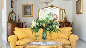 Een kamer met een geel gestreept bankje, een glazen vaas met bloemen, een kroonluchter en een spiegel met gouden lijst.