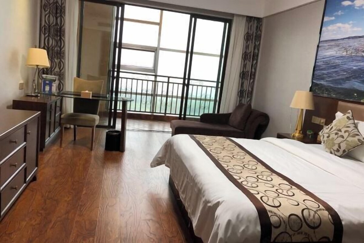 A hotel room with a bed, a desk, a chair, a balcony with a view, and a painting on the wall.