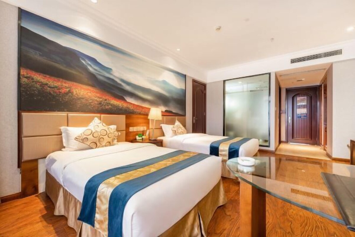 A hotel room with two beds, a glass table, a painting on the wall, and a door leading to another room.