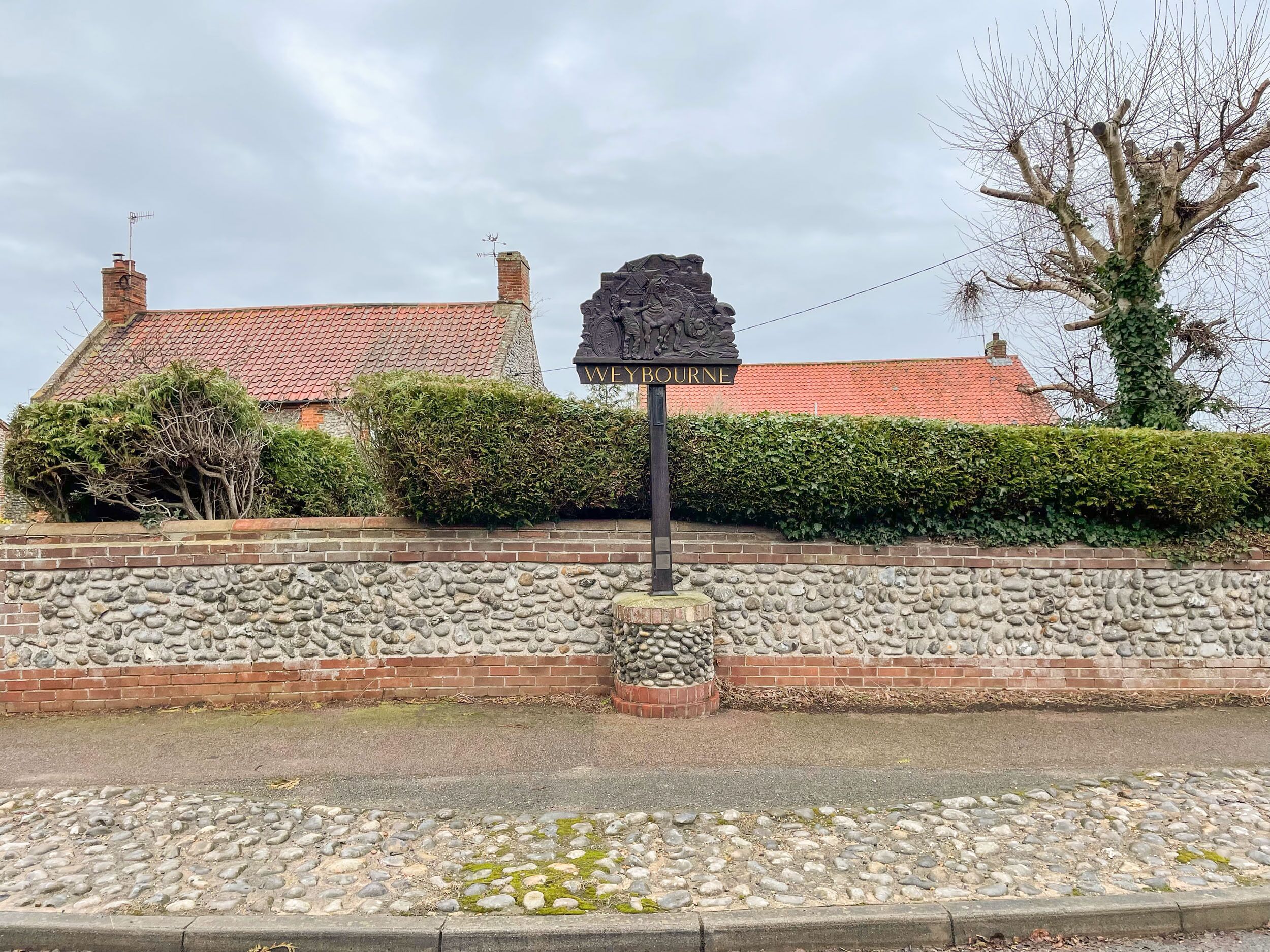 Un muro in pietra con un cartello 'Weybourne', un edificio in mattoni con tetto in tegole rosse e un siepe davanti.