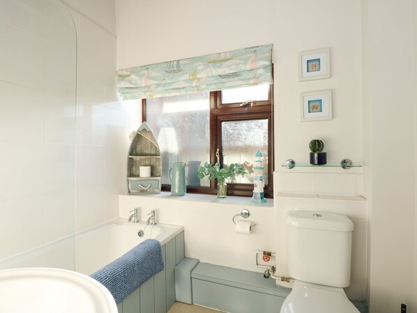 A bathroom with a bathtub, sink, and toilet. There is a window with a sailboat pattern curtain, a shelf with decorative items, and a small cactus plant. - Timbers (Holt)