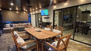 Un amplio espacio exterior con comedor, que incluye una mesa y sillas de madera, un sofá y un televisor montado en la pared.