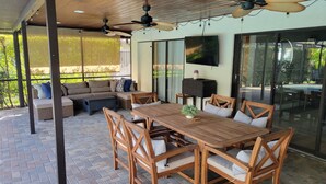 Un área de patio techada con una mesa y sillas de madera, un sofá y ventiladores de techo.