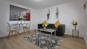 Una sala moderna con una mesa de centro de vidrio, un sofá negro con cojines amarillos, un comedor con una mesa y sillas blancas, y un cuadro de un paisaje urbano en la pared.