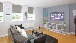 A modern living room with a sofa, a coffee table, a TV, and a shelving unit with decorative items. - Charming 2 Bed 2 Bath Flat & Parking by CozyNest (Reading)