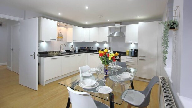 A modern kitchen with white cabinets, a black countertop, and a dining area with a glass table set for four. - Charming 2 Bed 2 Bath Flat & Parking by CozyNest (Reading)