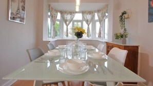 A dining room with a glass table set for six, white chairs, a vase with flowers, and a window with curtains.