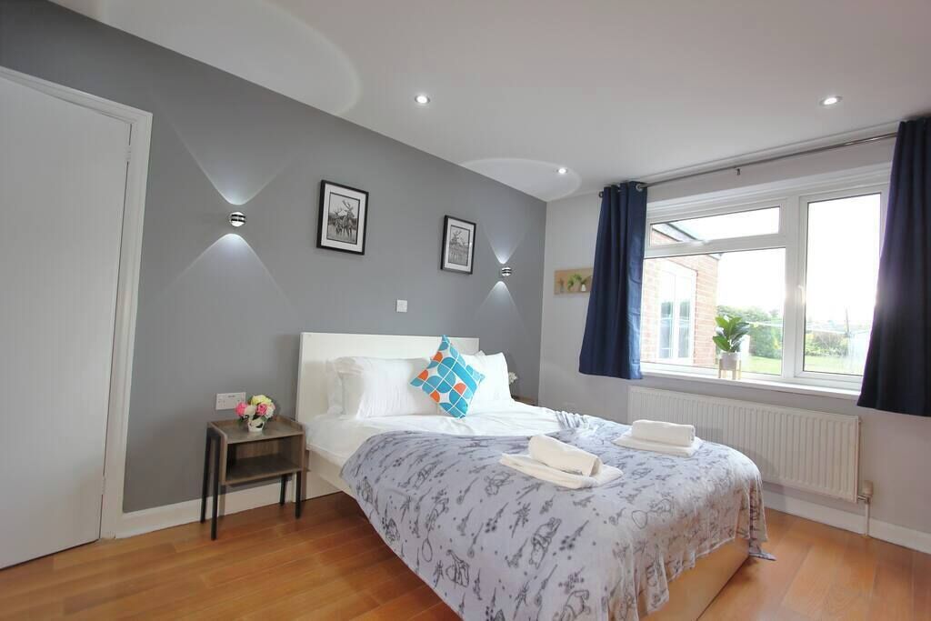 A bedroom with a bed, a bedside table with a vase, a window with curtains, and a wall-mounted light fixture.