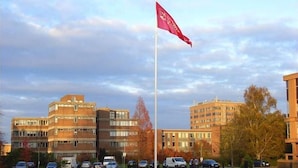 A flag on a flagpole in front of a building with multiple windows and a parking lot with cars.