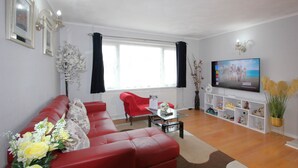 A living room with a red sofa, a red armchair, a coffee table, and a TV on the wall. There is a window with black curtains, a vase with flowers, and a shelf with decorative items.