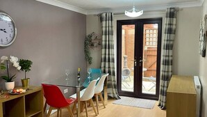A dining area with a glass table, red and white chairs, a clock on the wall, and a view of a wooden deck through a glass door. - Elegant 4 Bed 3 Bath House & Parking by CozyNest (Reading)