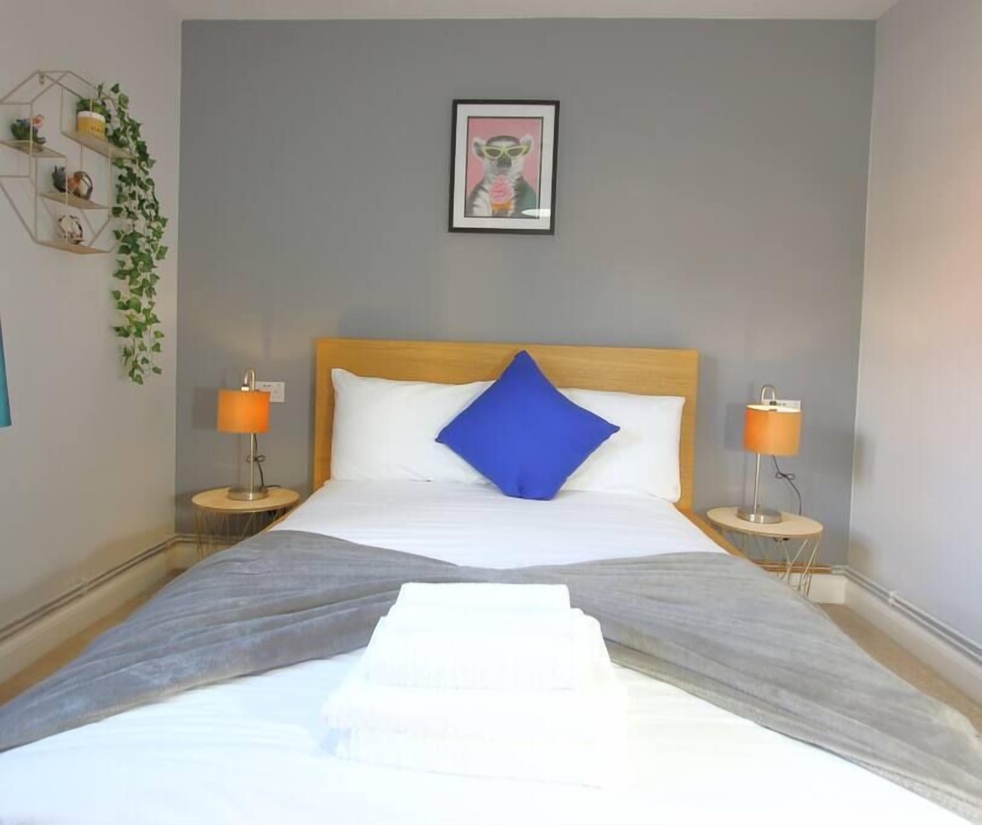 Habitación de hotel con cama, dos lámparas de noche, un cuadro en la pared y una planta pequeña a la izquierda.