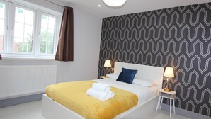 A modern hotel room with a large bed, two bedside tables with lamps, a patterned wall, and a window with curtains. - Elegant 4 Bed 3 Bath House & Parking by CozyNest (Reading)