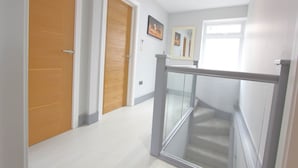 A hallway with a wooden door, a framed picture on the wall, and a staircase with a glass railing. - Elegant 4 Bed 3 Bath House & Parking by CozyNest (Reading)