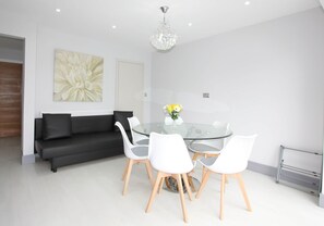 Un espace de salle à manger moderne avec une table en verre, des chaises blanches, un canapé noir, un arrangement floral en guise de décoration centrale et un lustre.