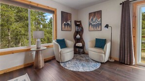 A cozy room with two leather chairs, a small round table with a lamp, a bookshelf, and a large window offering a view of trees.