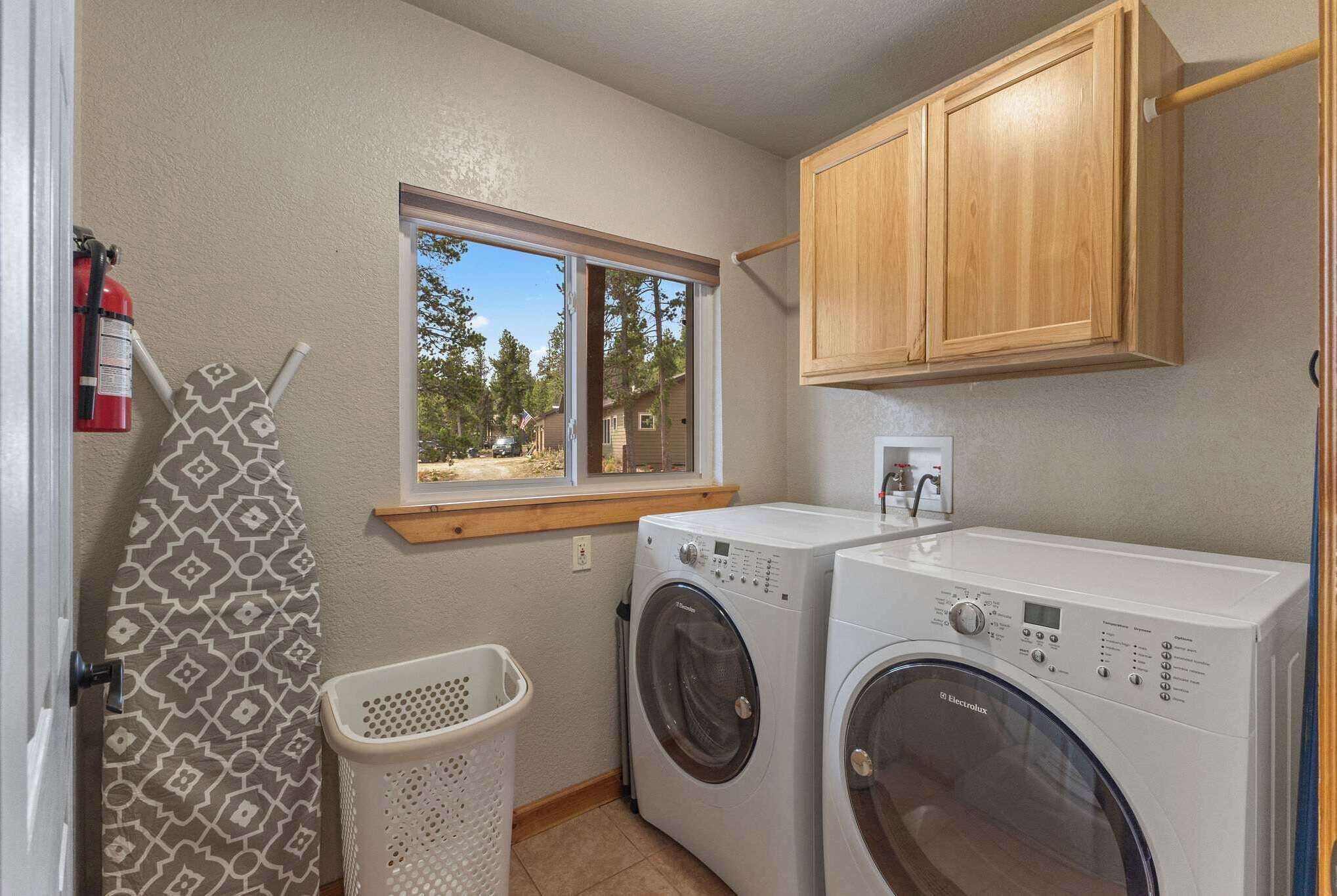 A laundry room with two front-loading washing machines, a drying rack, a window with a view of trees, and wooden cabinets.