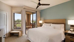 Quarto de hotel com uma cama grande, um ventilador de teto, uma escrivaninha com luminária e vista para o exterior.