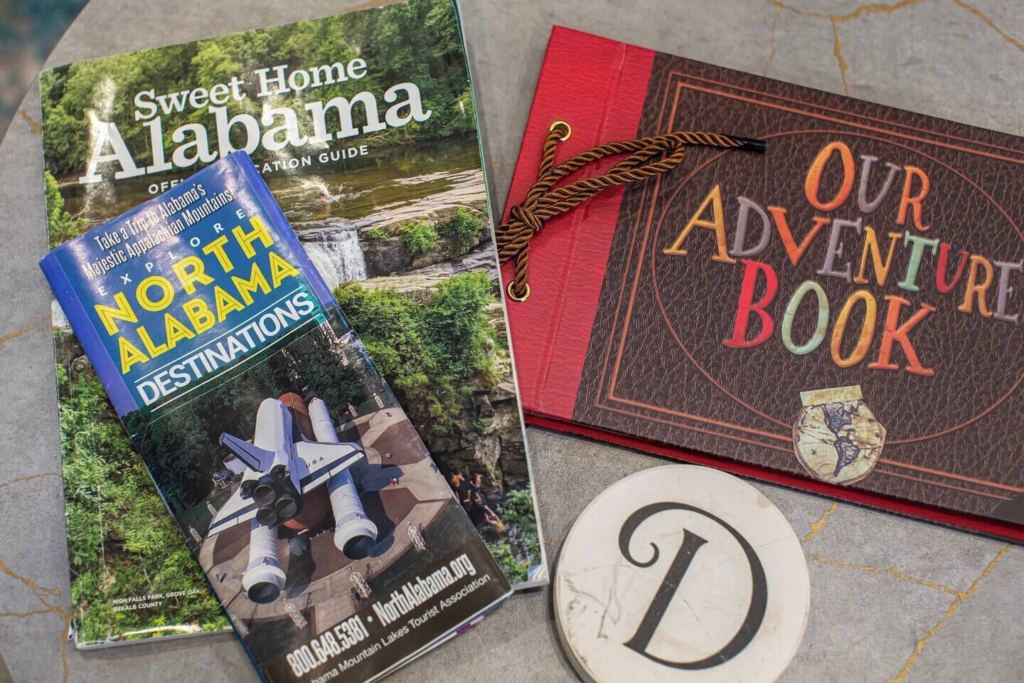 Zwei Reiseführer mit dem Titel 'Sweet Home Alabama' und 'Our Adventure Book', zusammen mit einem dekorativen Buchstaben 'D'.