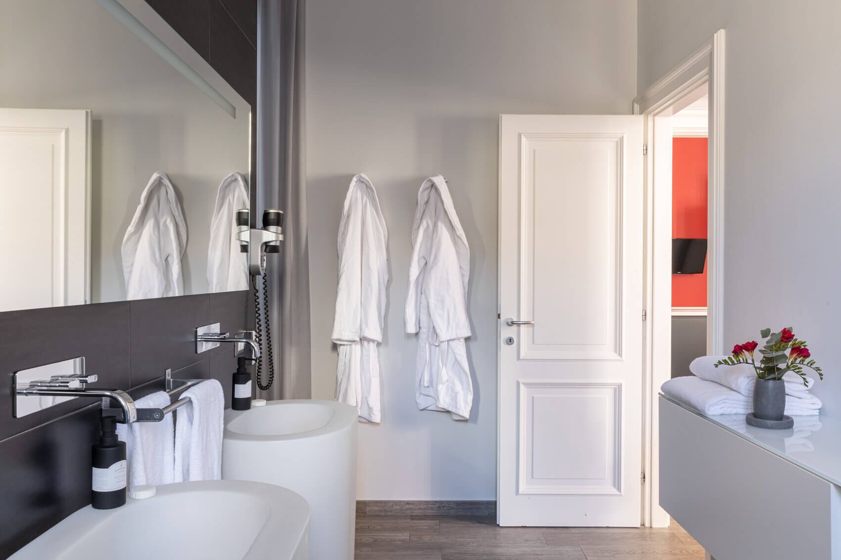 Un baño moderno con bañera, un espejo grande y zona de ducha. Hay toallas blancas y un florero con flores en la encimera.