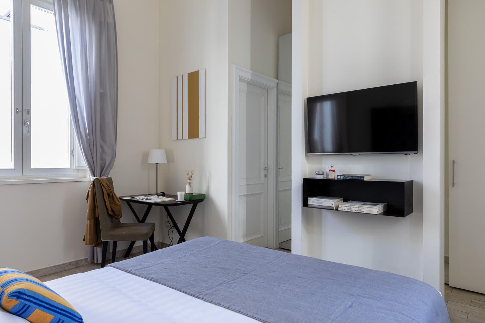 A hotel room with a bed, a desk with a lamp, a television mounted on the wall, and a door leading to another room.
