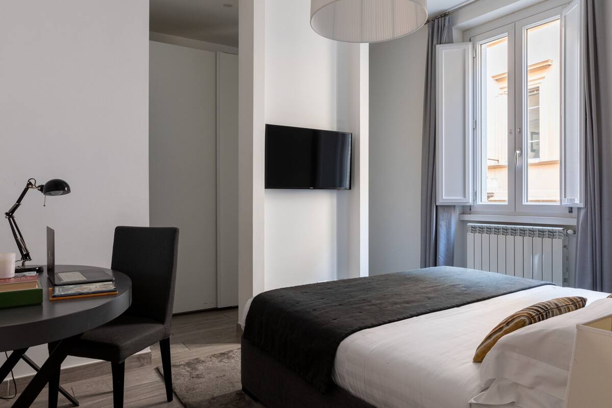 Une chambre d’hôtel moderne équipée d’un lit, d’un bureau avec un ordinateur portable, d’une chaise, d’un téléviseur fixé au mur et d’une fenêtre avec des rideaux.