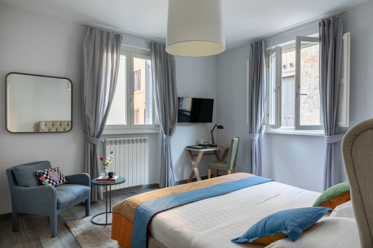 Une chambre d’hôtel avec un lit, une chaise, une petite table avec une lampe, un téléviseur et une fenêtre avec des rideaux.