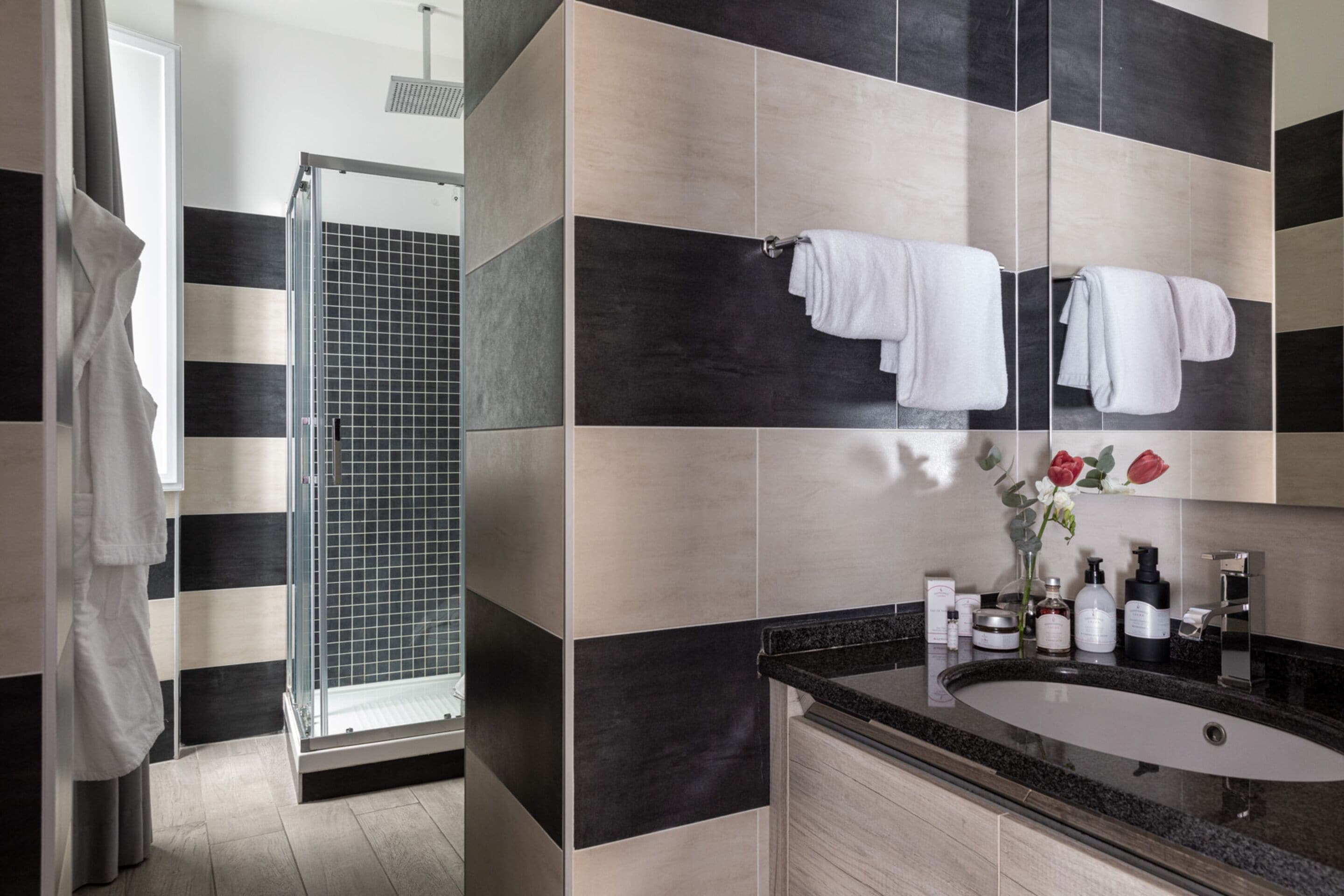 Un baño moderno con un espejo grande, baldosas negras y beige, un box de ducha de vidrio y un lavamanos con artículos de aseo.