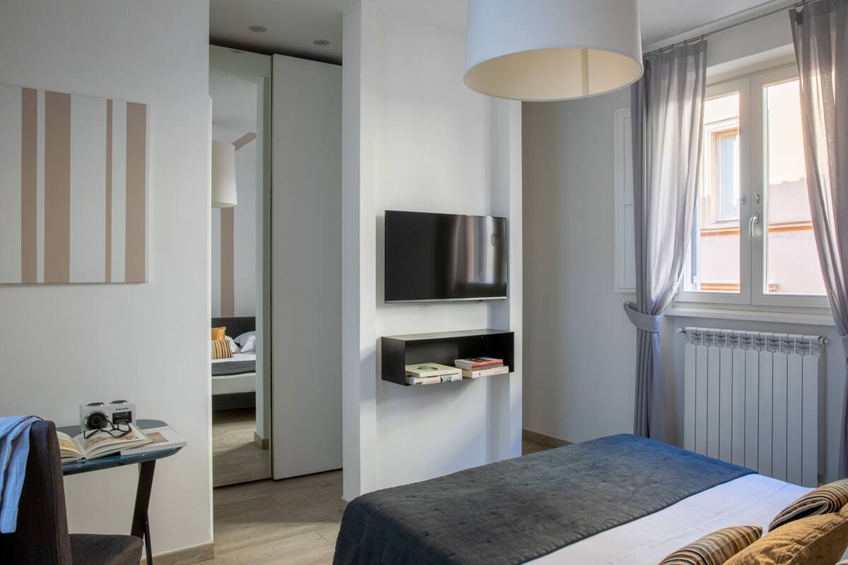 Une chambre d’hôtel moderne équipée d’un lit, d’un téléviseur fixé au mur, d’une petite table avec une chaise et d’une fenêtre avec des rideaux.