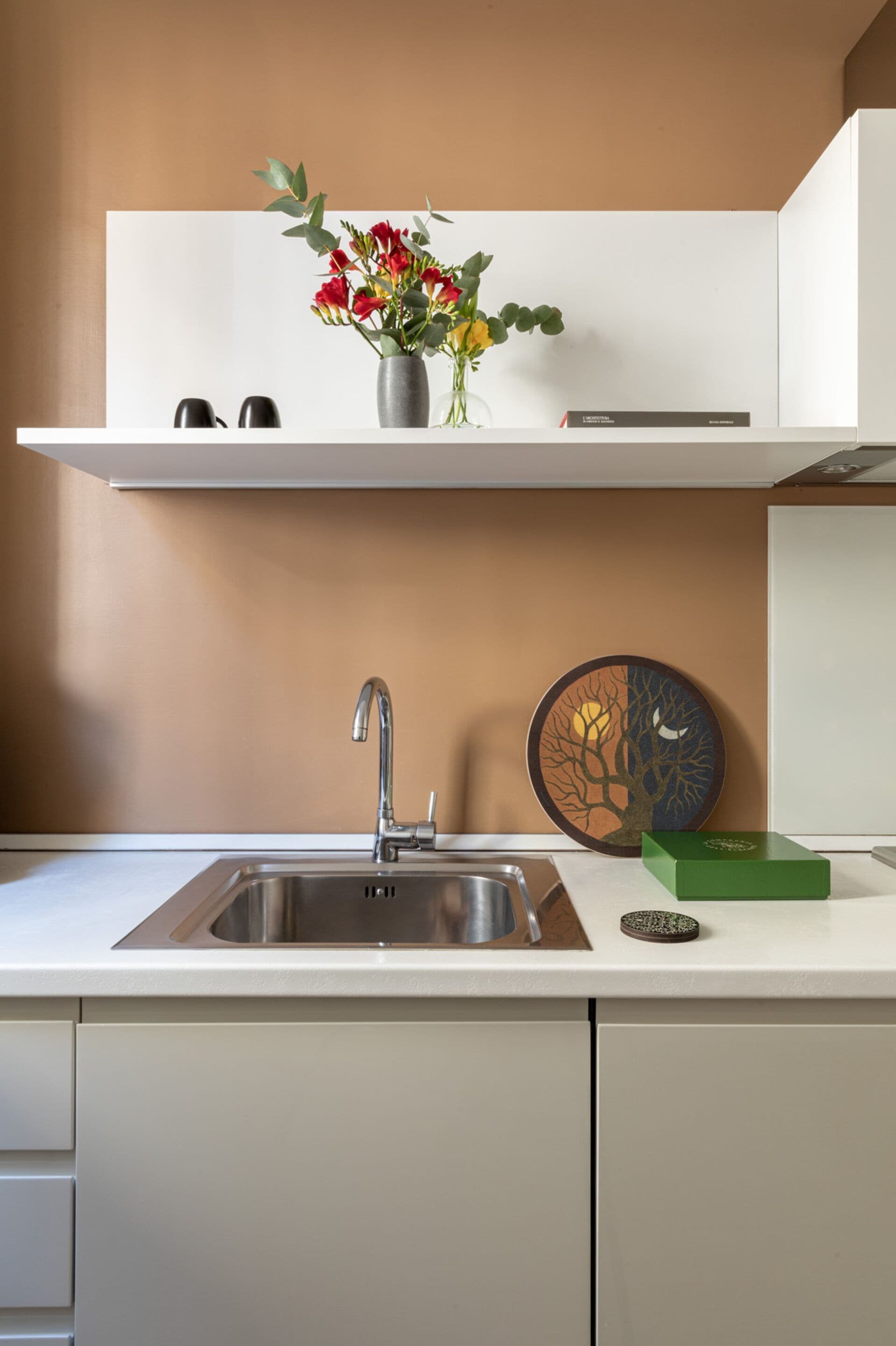 Una cucina con un lavandino in acciaio inossidabile, un vaso con fiori, un piatto decorativo e una scatola verde sul piano di lavoro.