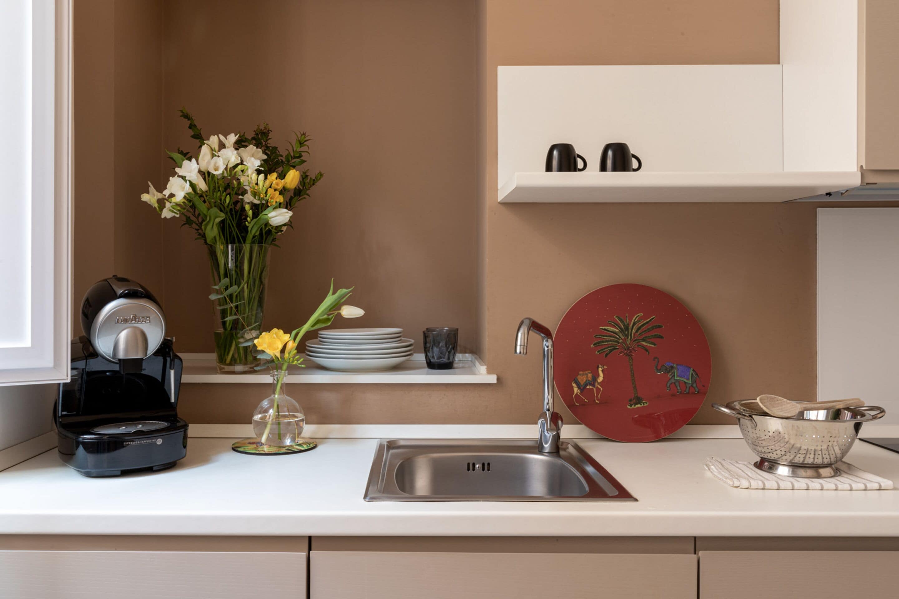 Una cocina con cafetera, un florero con flores, un lavamanos y una repisa con platos y una taza.