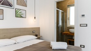 Une chambre d’hôtel moderne avec un grand lit, une table de chevet, une commode, une lampe fixée au mur, un tableau encadré, une salle de bain avec une douche en verre et un lavabo.