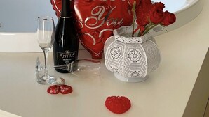 Un meuble-lavabo de salle de bain agrémenté d’un coussin en forme de cœur, une bouteille de Prosecco Antus, un verre, un vase rempli de roses et une assiette décorative.