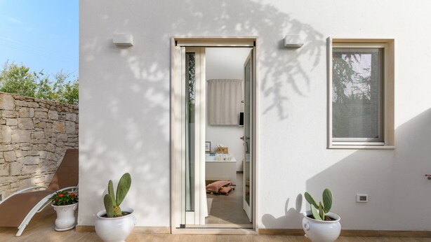 Une maison moderne avec un mur en pierre, une porte en verre donnant accès à une salle de bain et deux cactus en pot.