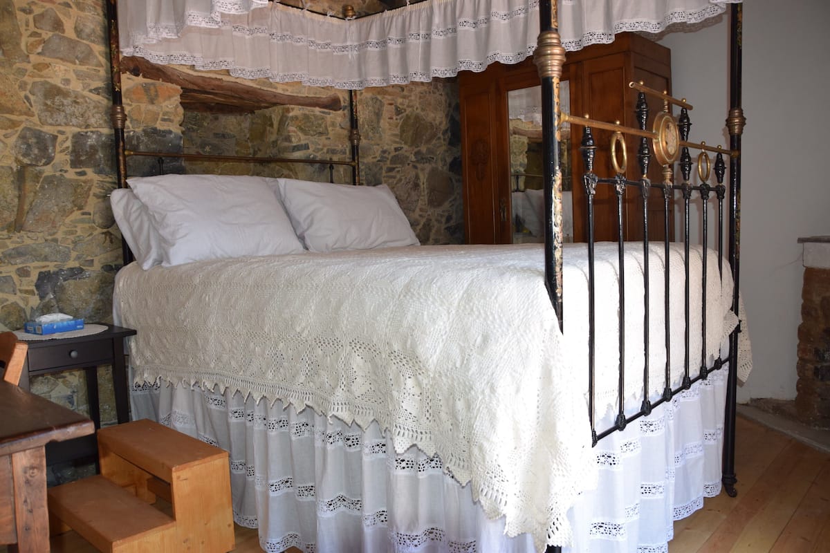 Una cama con dosel, con cubrecama blanco y encaje, frente a una pared de piedra y una puerta de madera al fondo.